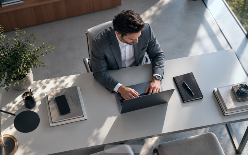 Man working on a laptop at a modern office desk, viewed from above, with notebooks, a phone and plants arranged around him.